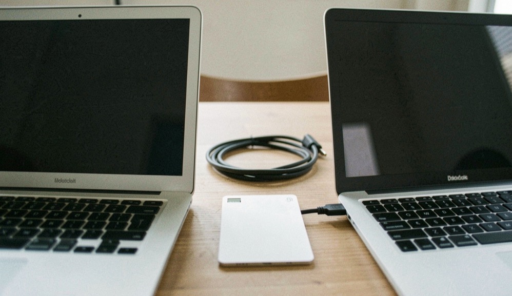 Documentary photograph of two different laptops beside a shared blank data card and a cable, screens turned away, no logos or writing, neutral daylight. Distinct home journal 3 composition for this editorial entry.