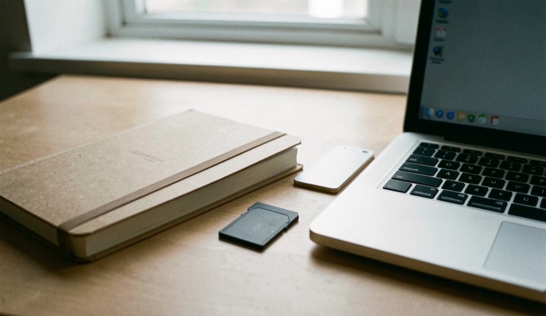 Documentary photograph of a notebook, export folder and blank memory card beside a laptop, no readable screen or writing, quiet window light. Distinct home journal 6 composition for this editorial entry.